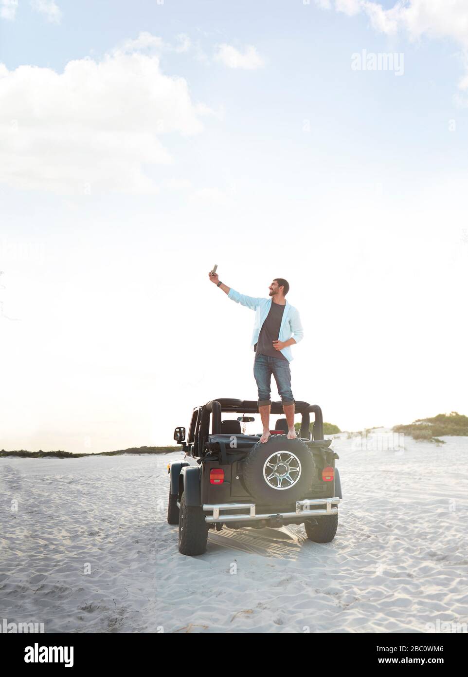 Junger Mann mit Kameratelefon, der selfie auf Jeep-Reifen am Strand nimmt Stockfoto