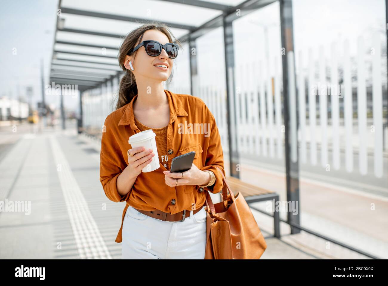 Lifestyle-Porträt einer jungen und fröhlichen Frau, die mit Telefon und Kaffeetasse auf den öffentlichen Verkehrsmitteln steht, hält im Freien. Städtisches Geschäftsreise-Konzept Stockfoto