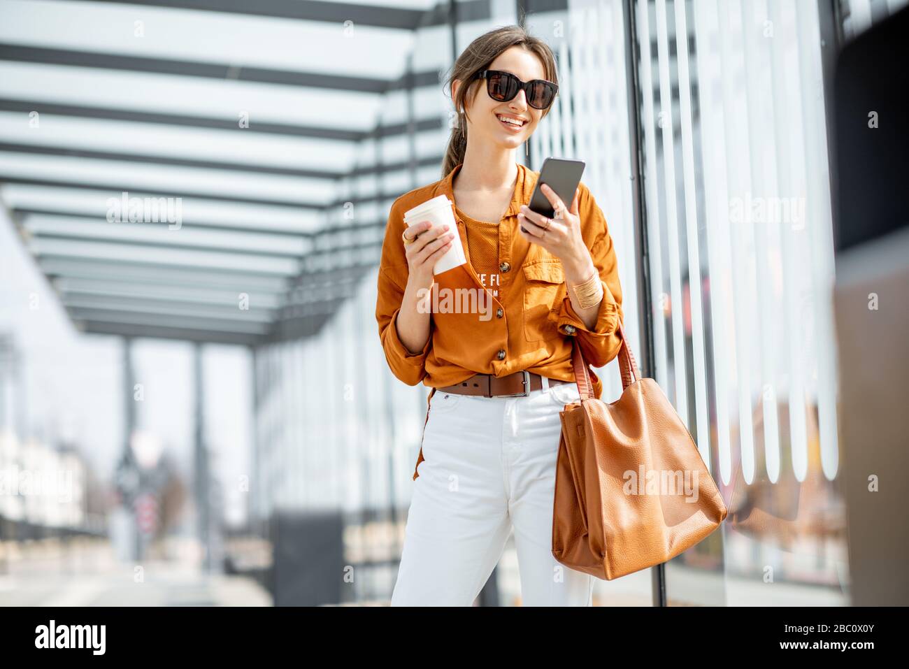 Lifestyle-Porträt einer jungen und fröhlichen Frau, die mit Telefon und Kaffeetasse auf den öffentlichen Verkehrsmitteln steht, hält im Freien. Städtisches Geschäftsreise-Konzept Stockfoto