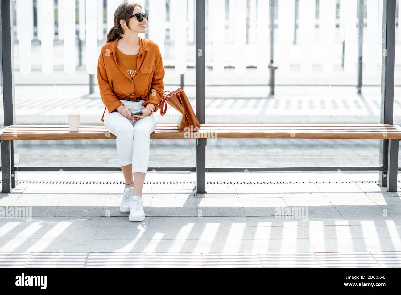 Eine fröhliche, stilvolle Frau, die mit Telefon und Tasche an einem modernen öffentlichen Verkehrsmittel sitzt, hält an einem sonnigen Tag im Freien. Konzept eines städtischen Verkehrs und urbun Life Stockfoto