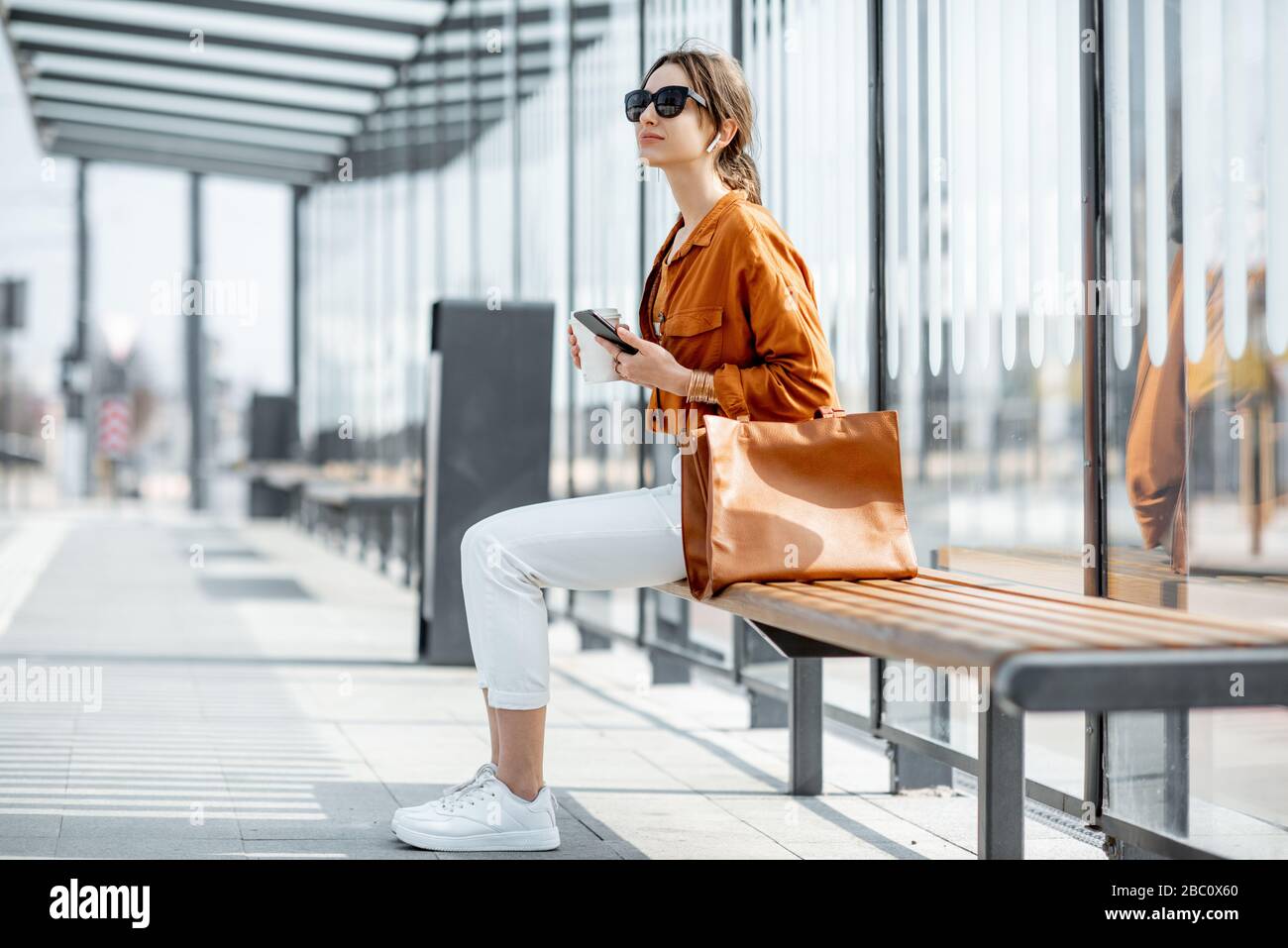 Junge, stilvolle Frau, die auf die öffentlichen Verkehrsmittel wartet und mit Telefon und Kaffee an der Haltestelle draußen sitzt. Städtisches Geschäftsreise-Konzept Stockfoto