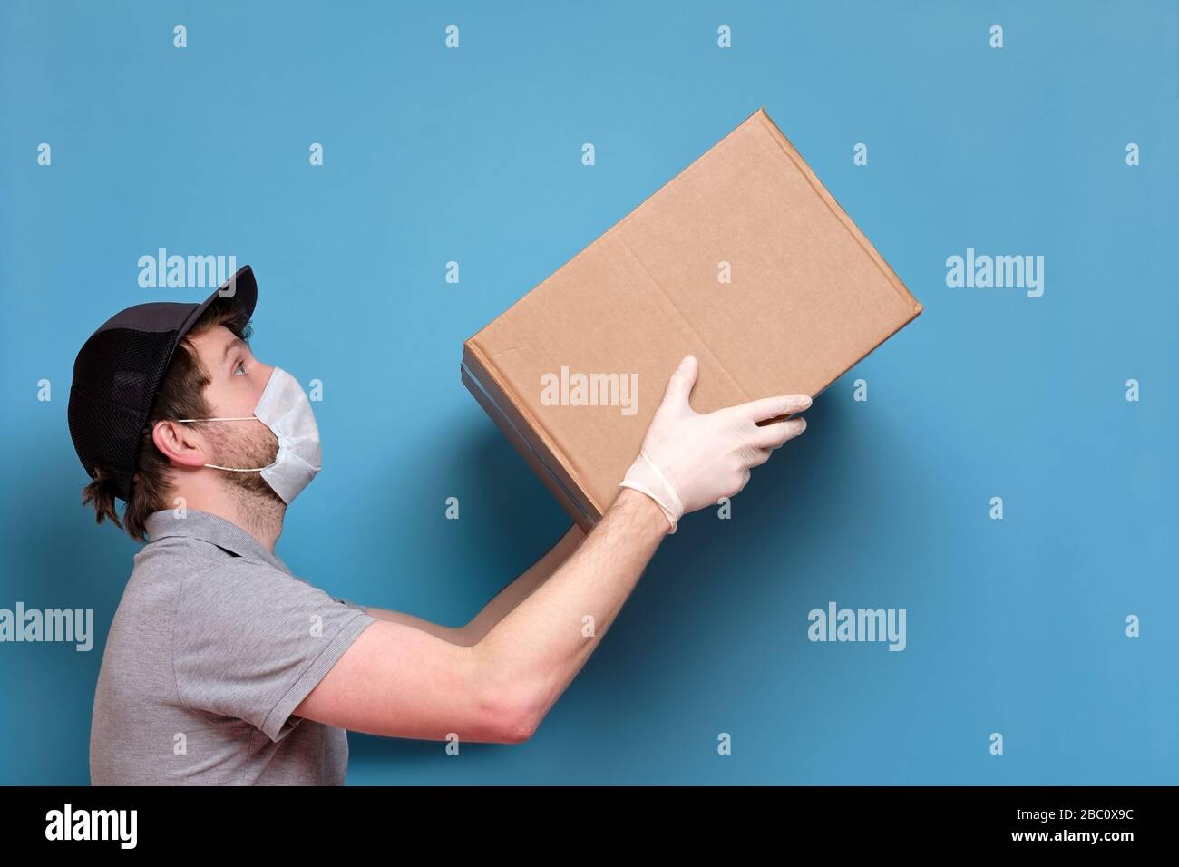 Kaukasischer junger Mann in medizinischer Maske, der ein Paket hält, das er dem Kunden gibt. Kein ansteckendes Lieferkonzept. Studio an blauer Wand gedreht. Stockfoto