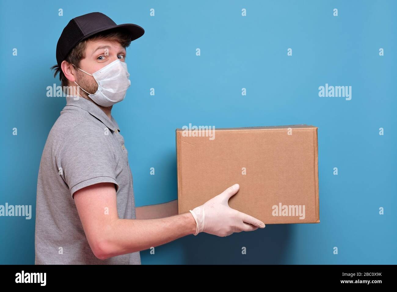 Kaukasischer junger Mann in medizinischer Maske, der ein Paket hält, das er dem Kunden gibt. Kein ansteckendes Lieferkonzept. Studio an blauer Wand gedreht. Stockfoto