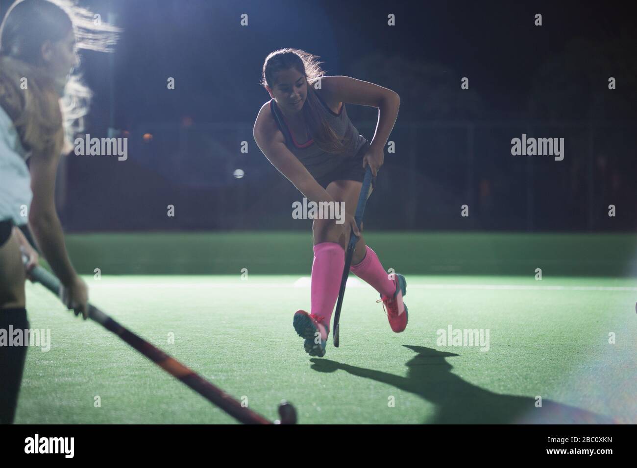 Entschlossener junger Hockeyspieler, der mit Hockeyschläger läuft und nachts auf dem Feld spielt Stockfoto