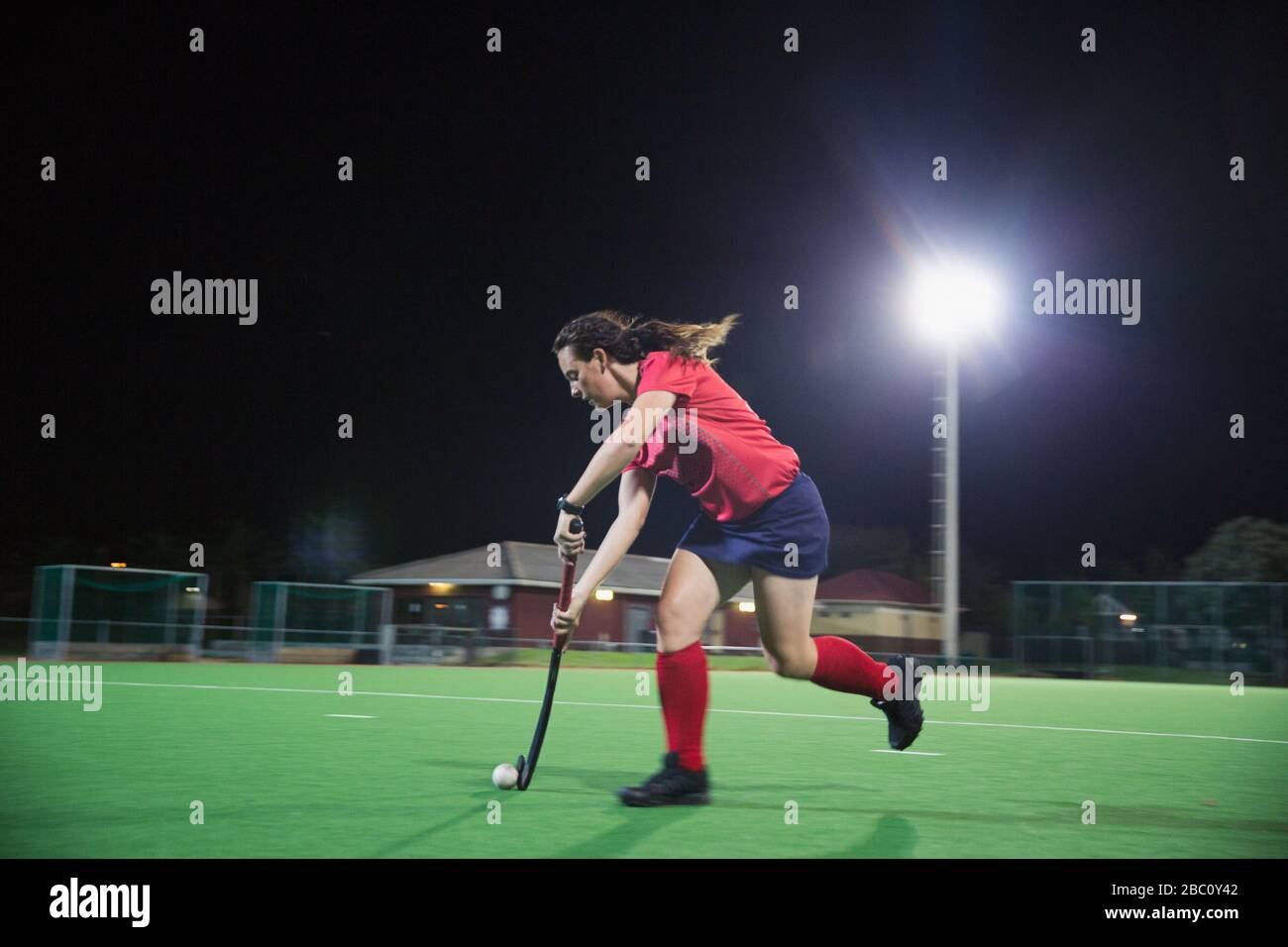 Entschlossener junger Hockeyspieler, der mit Hockeyschläger und Ball läuft und nachts auf dem Feld spielt Stockfoto