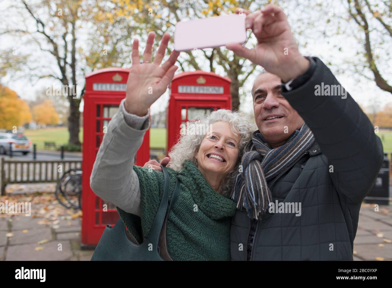 Lächelndes Senioren-Paar nimmt selfie im Herbstpark vor roten Telefonzellen Stockfoto