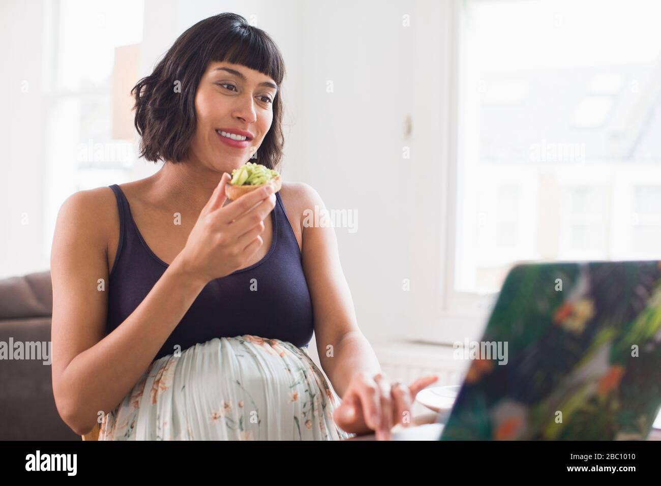 Glückliche schwangere Frau, die Avocado Toast auf dem Laptop isst Stockfoto