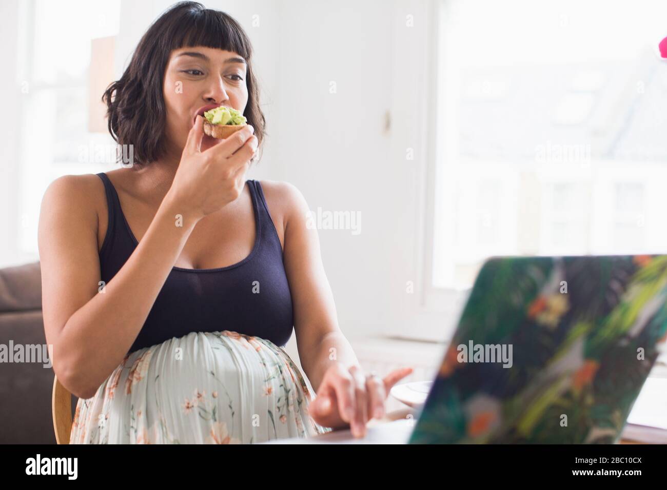 Schwangere Frau, die Avocado Toast auf dem Laptop isst Stockfoto