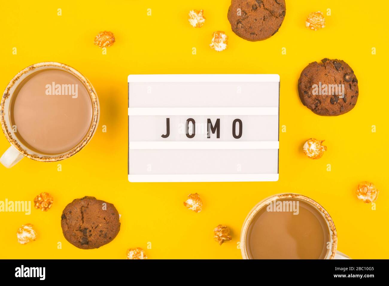 Kürzel Wort JOMO auf einer dekorativen Tafel auf hellgelbem Hintergrund. Das Konzept der Entspannung durch Informationen und Geräte. Draufsicht, flacher Lay. Stockfoto