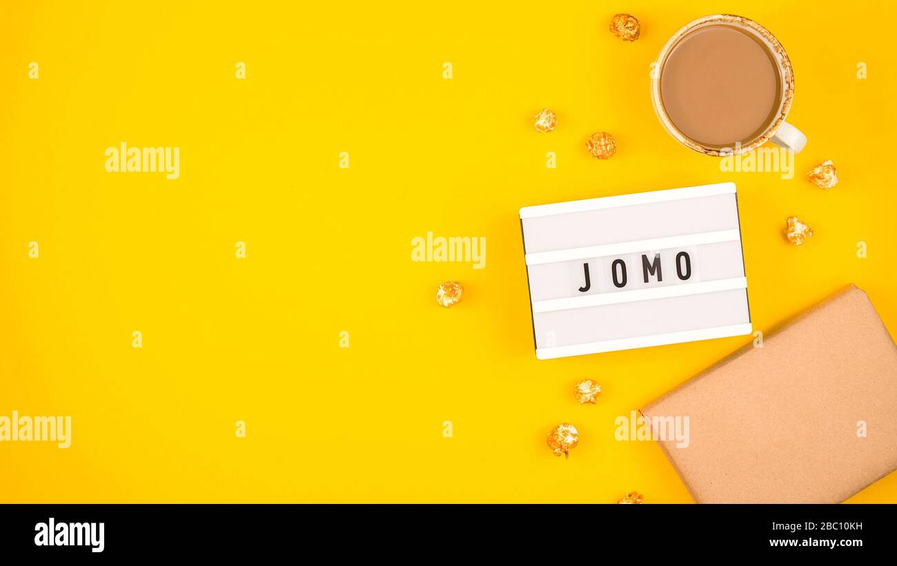 Kürzel Wort JOMO auf einer dekorativen Tafel auf hellgelbem Hintergrund. Das Konzept der Entspannung durch Informationen und Geräte. Draufsicht, flaches Layout, Kopierbereich. Stockfoto
