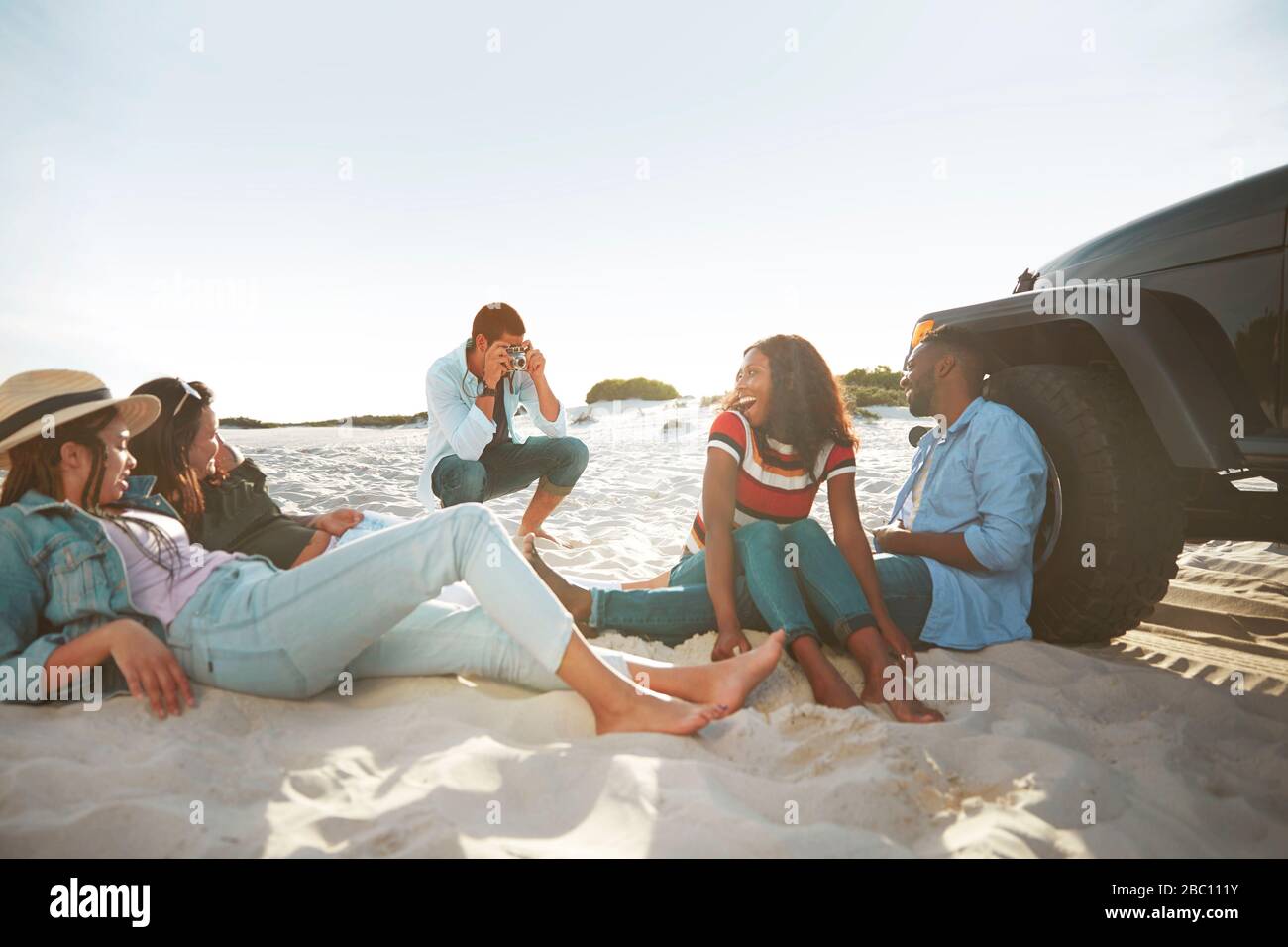 Junger Mann mit Digitalkamera fotografiert Freunde, die am sonnigen Strand hängen Stockfoto