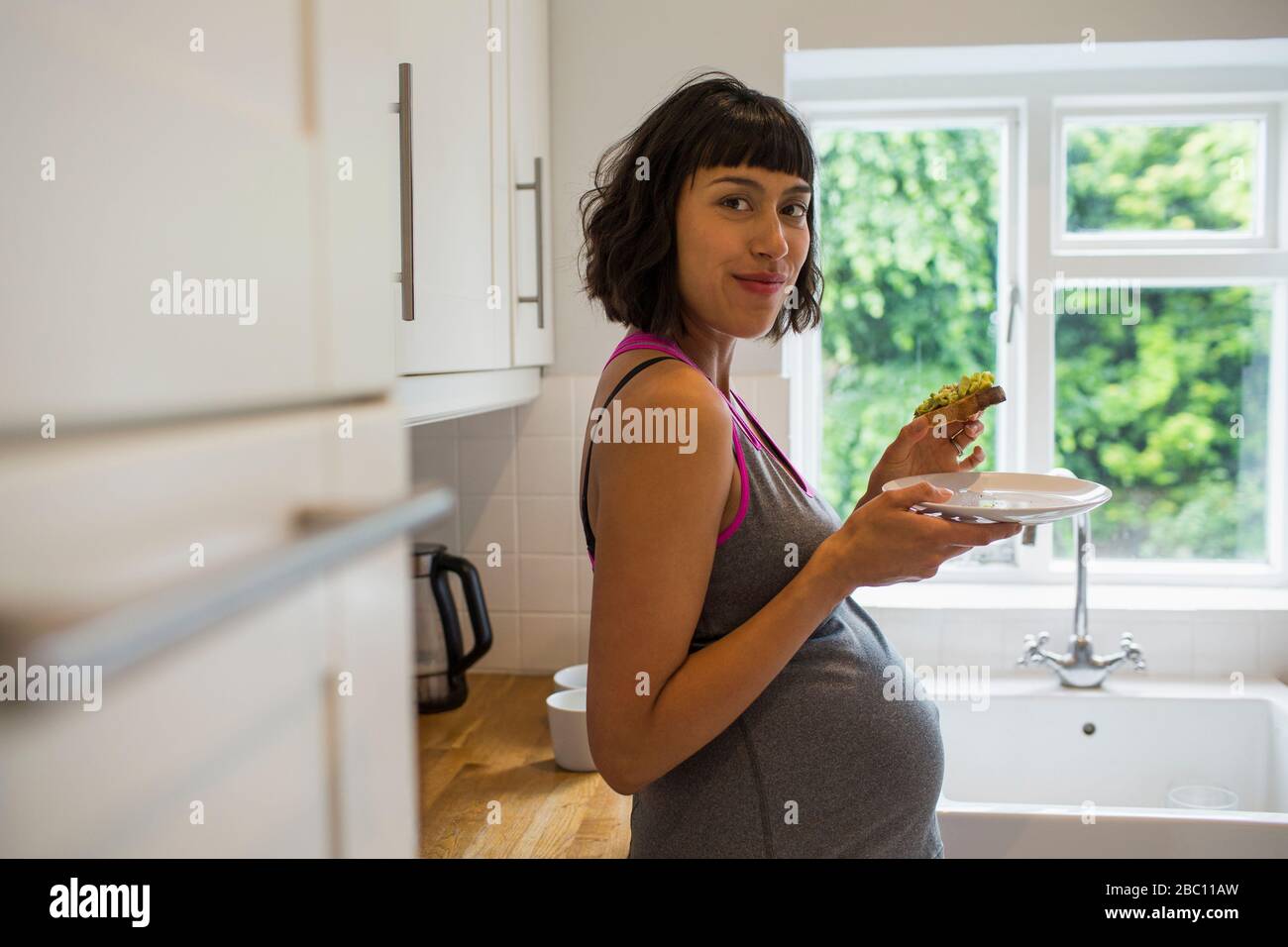 Portrait selbstbewusste schwangere Frau, die Avocado Toast in der Küche isst Stockfoto