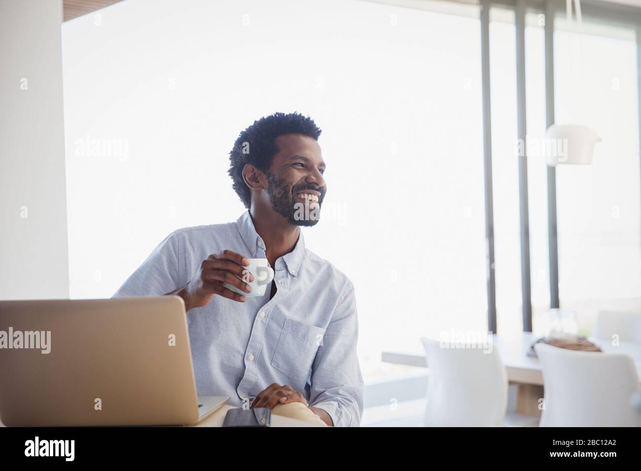 Lächelnder, begeisterter Mann, der Kaffee trinkt und zu Hause am Laptop arbeitet Stockfoto