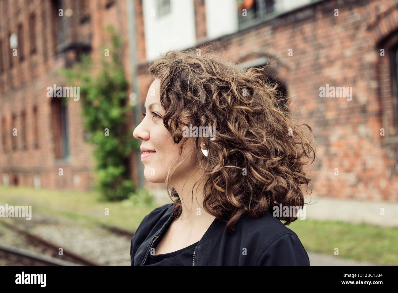 Lächelnde Brünette Frau mit lockigen Haaren vor einem Ziegelgebäude und alten Schienen Stockfoto