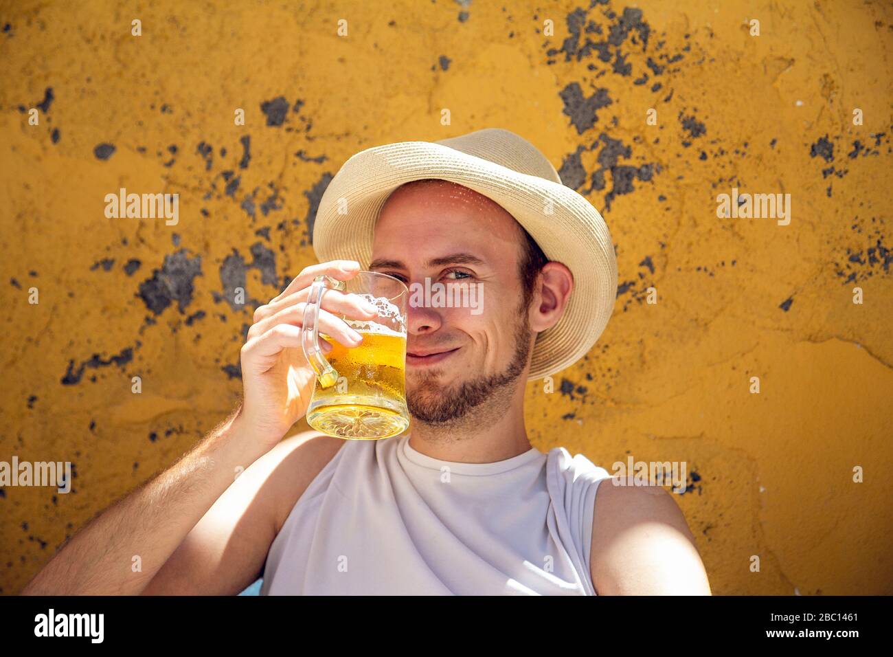 Junger Mann sitzt vor der abblätternden gelben Wand, trinkt Bier, rösten Stockfoto