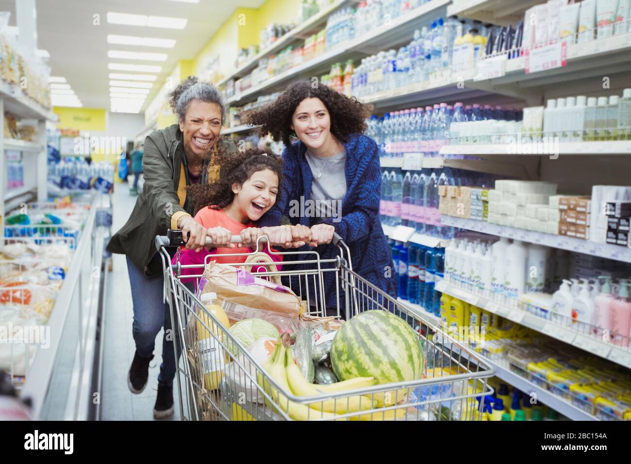 Verspielte Mehrgenerationsfrauen schieben Einkaufswagen in Supermarkt-Gang Stockfoto