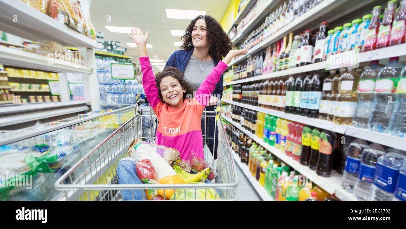 Mutter schiebt verspielte Tochter in den Einkaufswagen im Supermarktgang Stockfoto