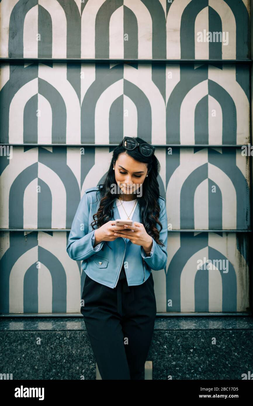 Porträt einer jungen Frau mit langen schwarzen Haaren, die mit dem Smartphone vor der gemusterten Wand steht Stockfoto