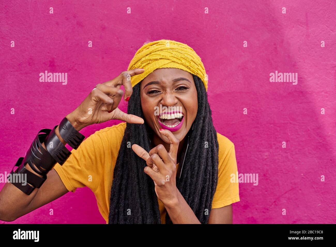 Porträt einer Frau mit langen Dreadlocks, die vorgibt, mit den Händen vor einer rosa Wand ein Foto zu machen Stockfoto
