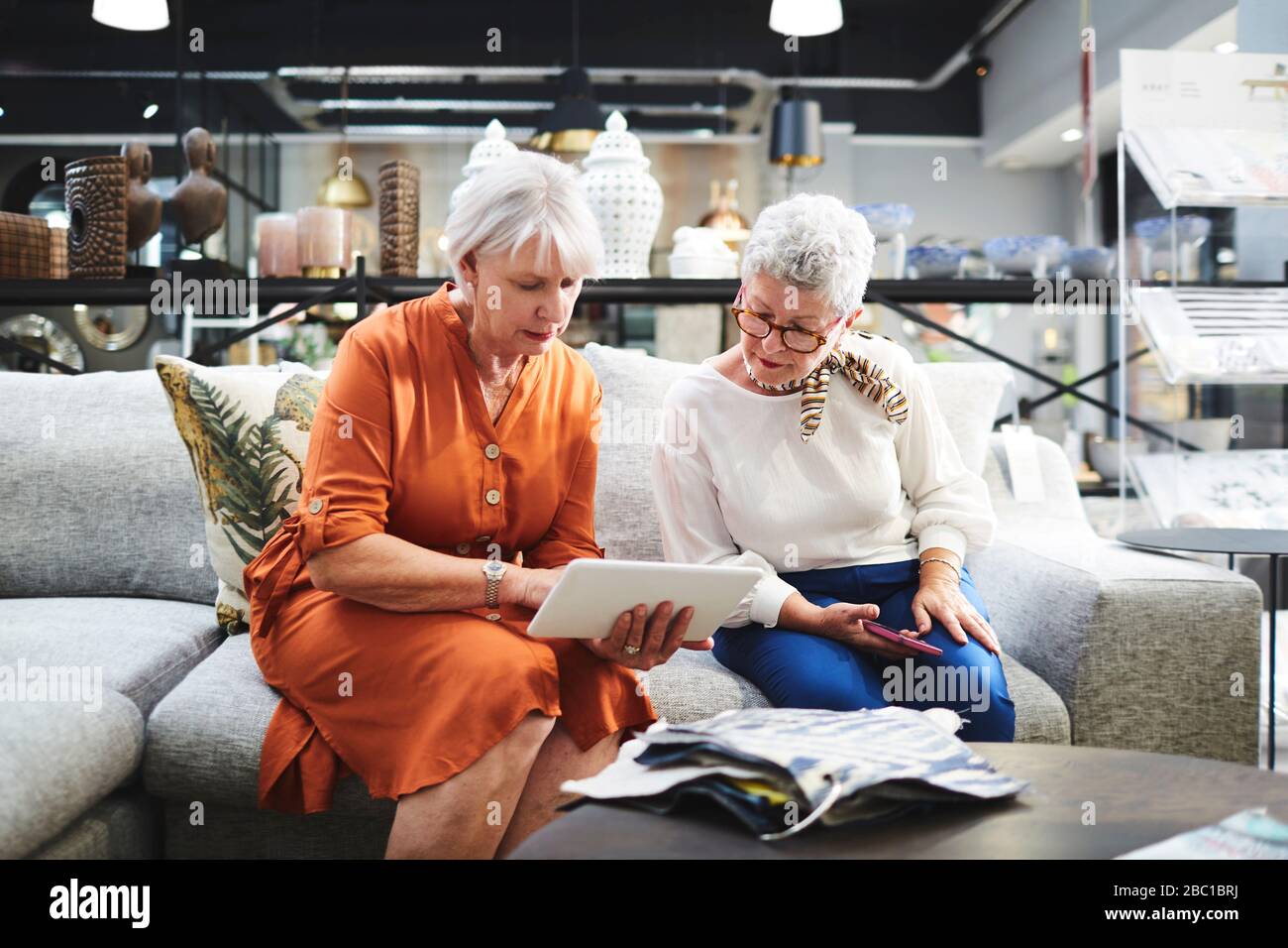 Seniorinnen mit digitalem Tablet, die Stoffschwatzen auf dem Sofa im Möbelgeschäft betrachten Stockfoto