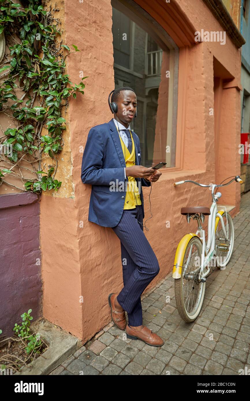 Stilvoller junger Geschäftsmann mit Fahrrad in altmodischem Anzug, der Musik auf seinen Kopfhörern hört Stockfoto