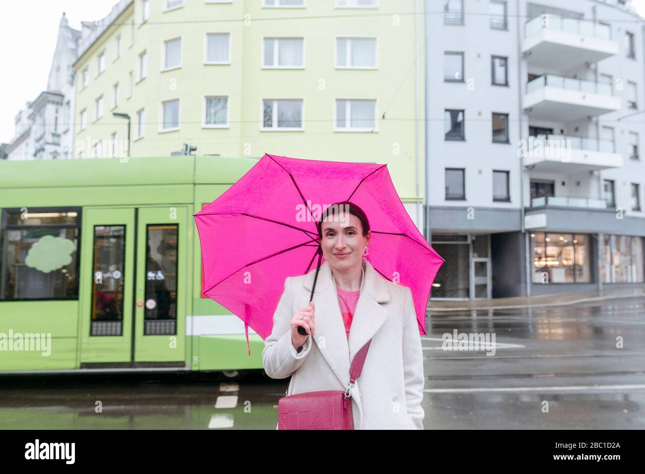 Lächelnde Frau mit rosa Regenschirm auf der Straße, Straßenbahn im Hintergrund Stockfoto
