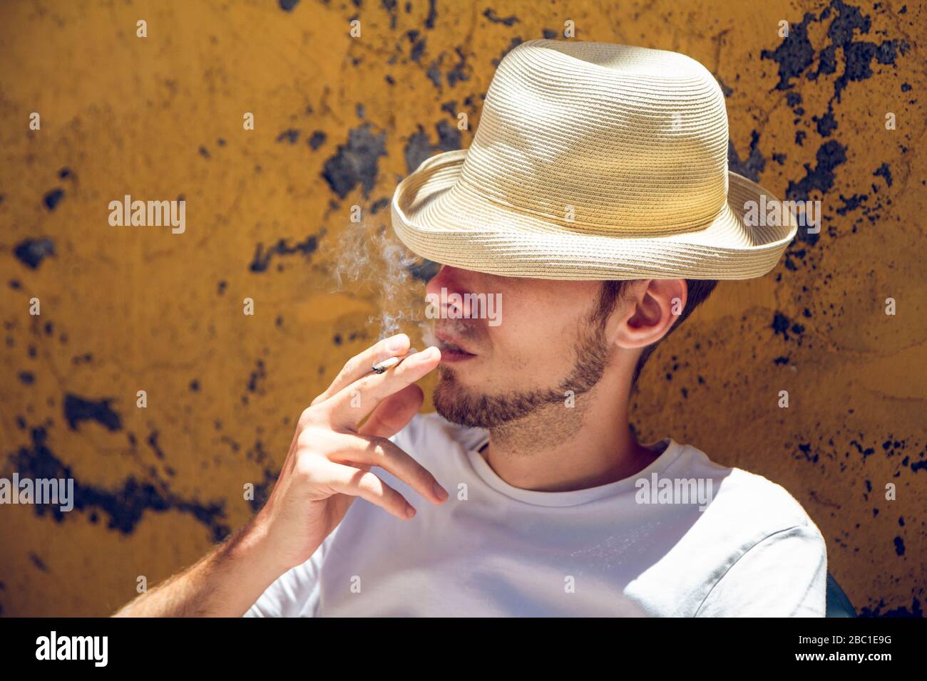 Junger Mann, der vor der abblätternden gelben Wand sitzt und eine Zigarette raucht Stockfoto