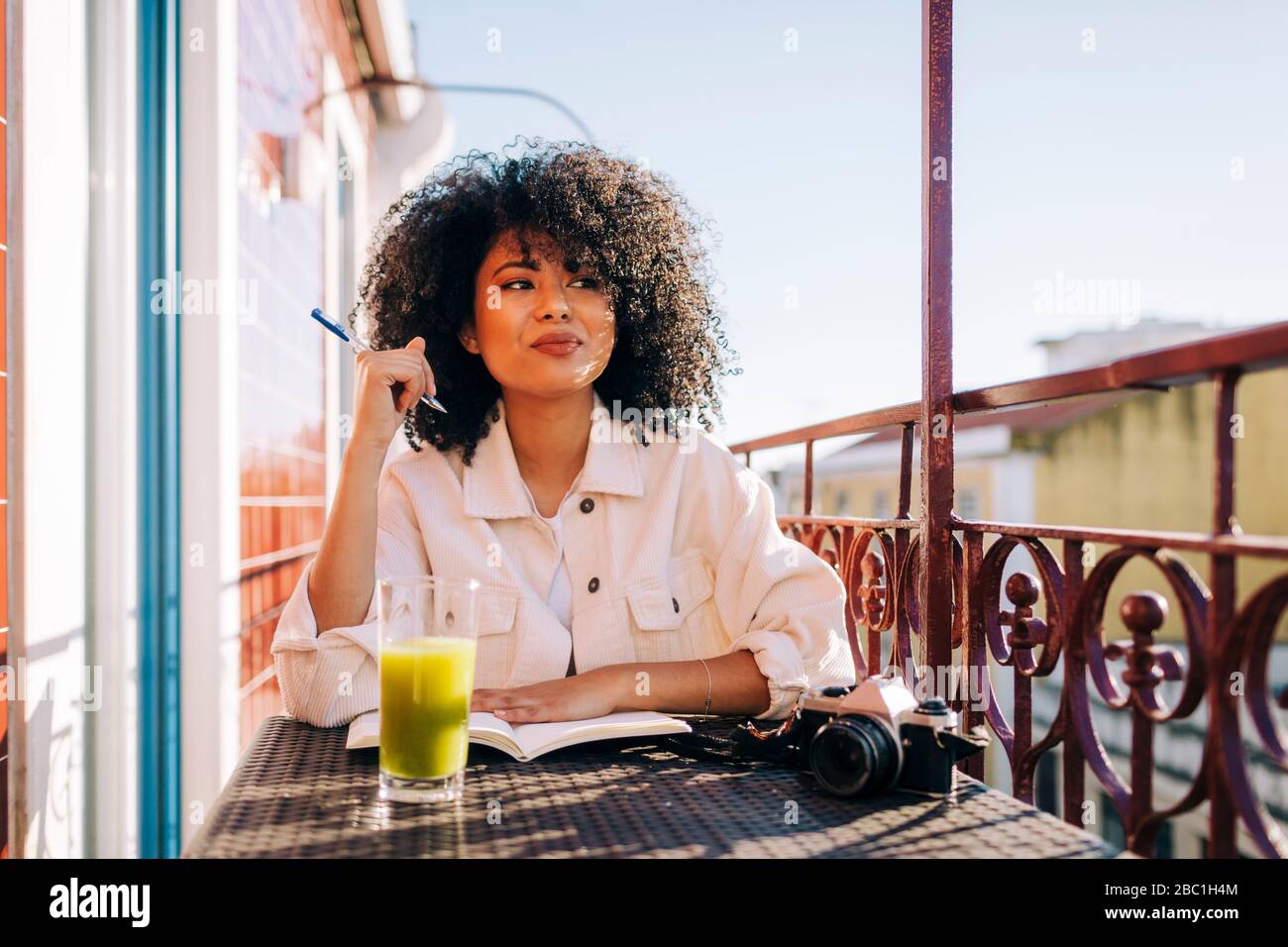 Lächelnde junge Frau mit lockigen Haaren sitzt am Tisch auf dem Balkon mit Notebook, Smoothie und Kamera Stockfoto