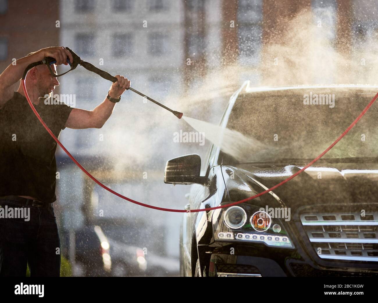 Seitenansicht des Mannes, der sein schwarzes Auto im Freien putzt, Wasser spritzt über die Oberseite des Autos, das auf die Sonne scheint. Stockfoto