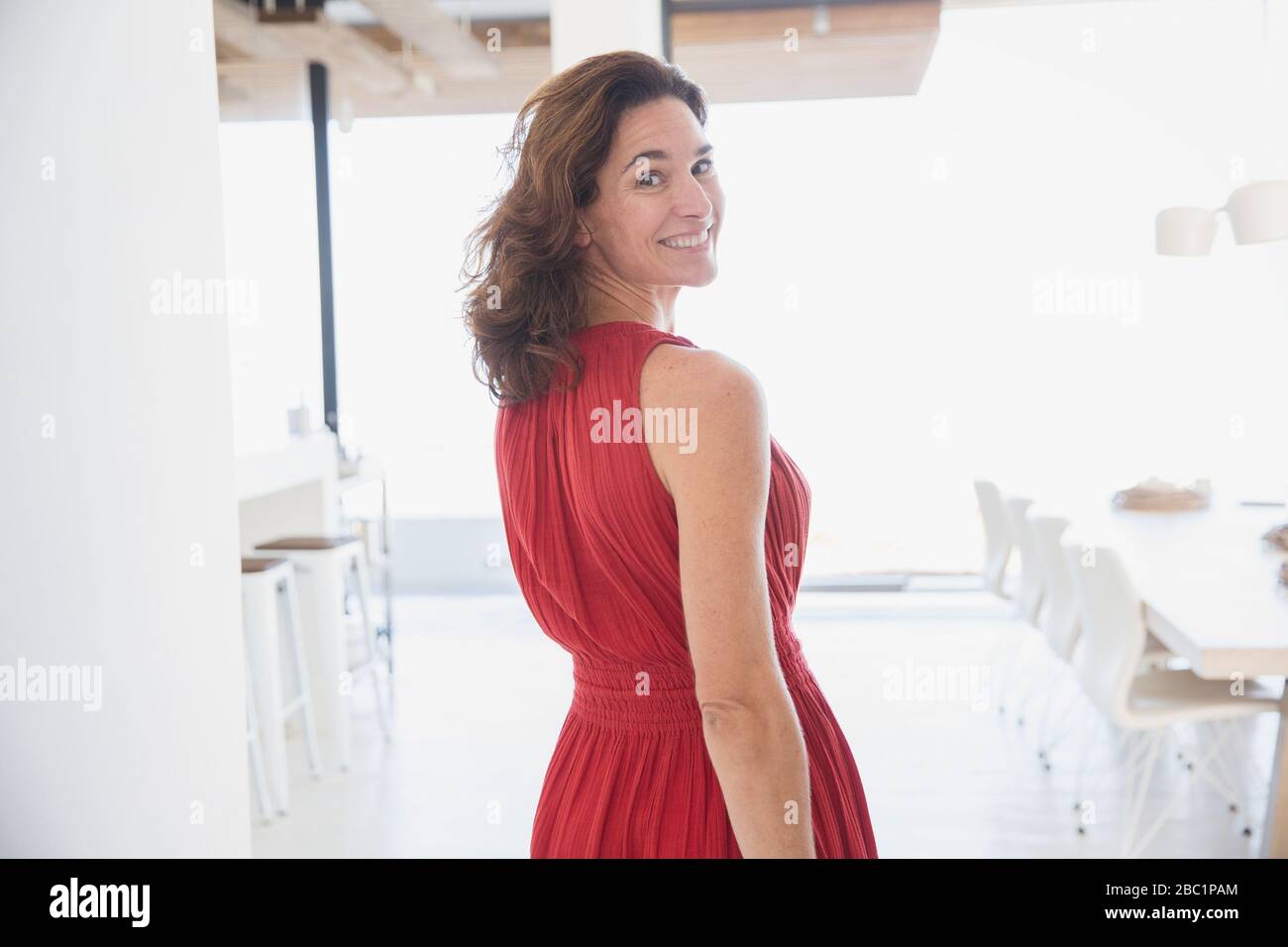 Portrait lächelnde, selbstbewusste brünette Frau im roten Kleid, die zu Hause über die Schulter blickt Stockfoto