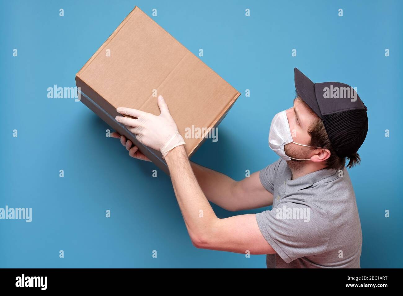 Kaukasischer junger Mann in medizinischer Maske, der ein Paket hält, das er dem Kunden gibt. Kein ansteckendes Lieferkonzept. Studio an blauer Wand gedreht. Stockfoto