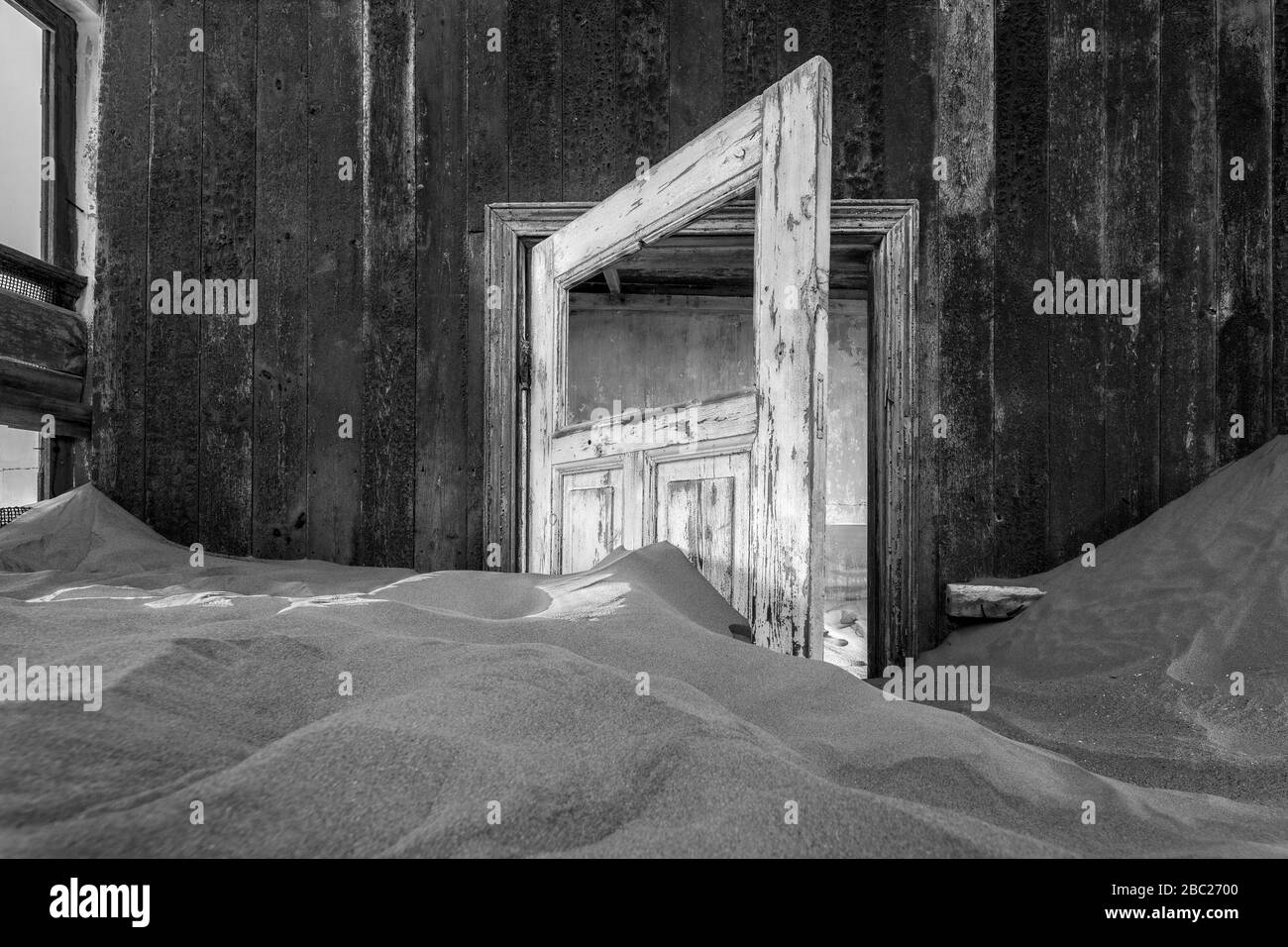 Ein Schwarz-Weiß-Foto in einem verlassenen Haus mit einer offenen Tür, die in dem gewellten Wüstensand untertaucht ist, aufgenommen in der Geisterstadt Kolmanskop, Stockfoto