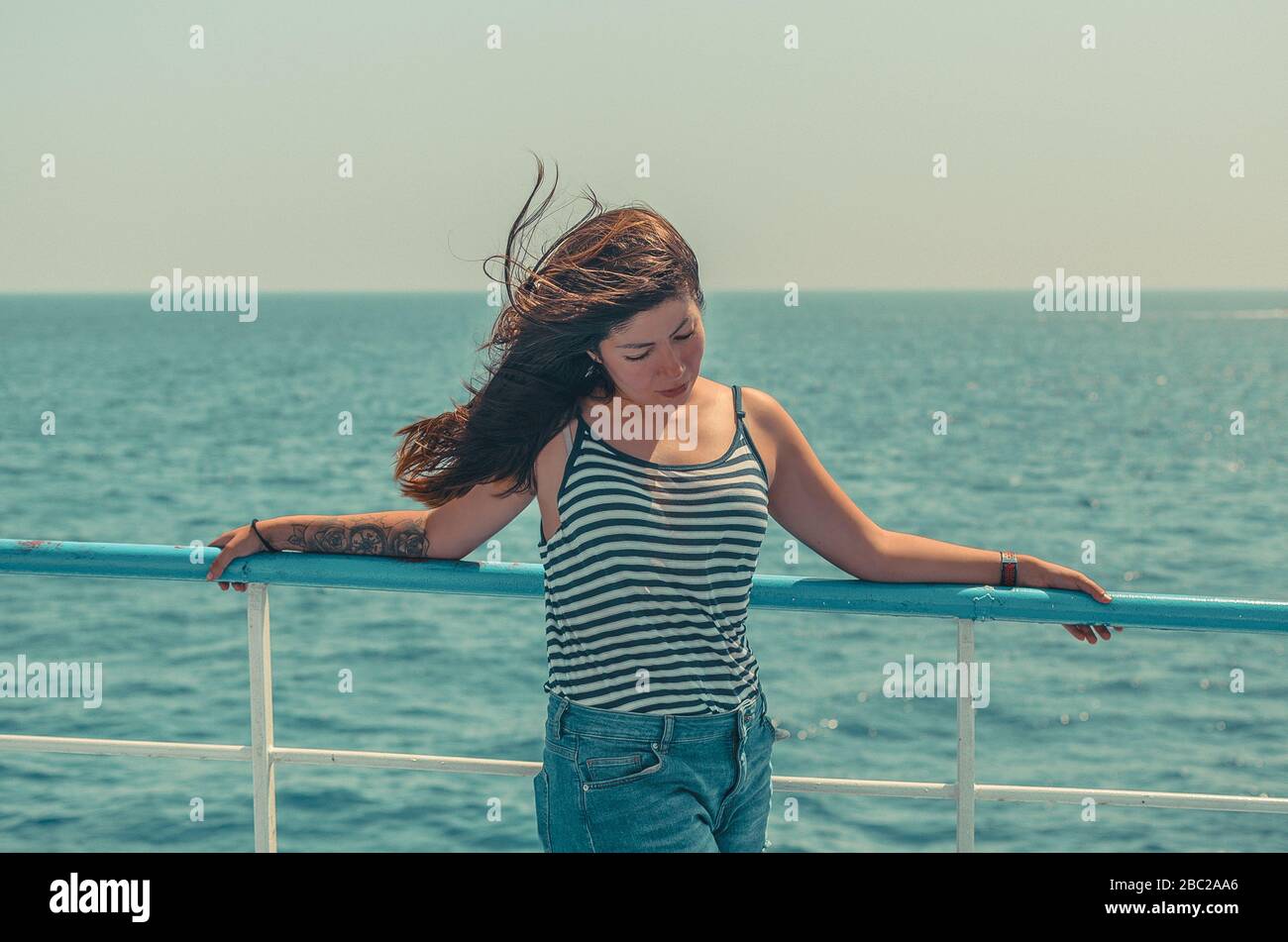 Hübsche junge brünette Frau mit flatternden Haaren in einem gestreiften T-Shirt und blauen Denim-Shorts genießt das Meer auf dem Deck eines Schiffes Stockfoto
