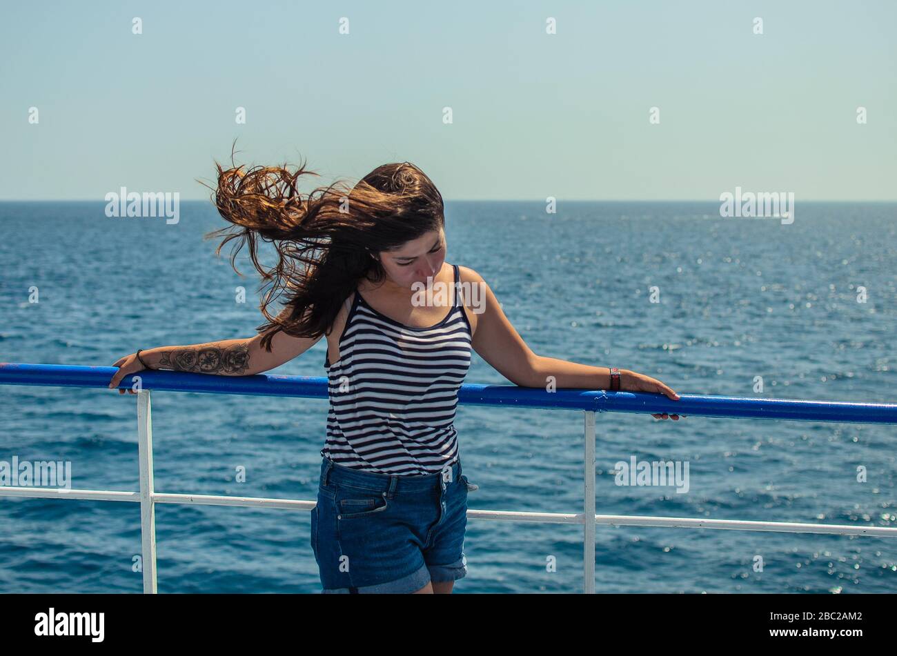 Hübsche junge brünette Frau mit flatternden Haaren in einem gestreiften T-Shirt und blauen Denim-Shorts genießt das Meer auf dem Deck eines Schiffes Stockfoto