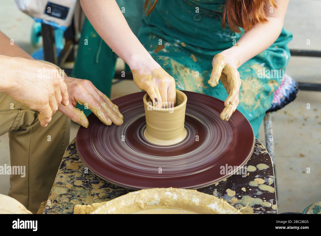 Die Hände eines Meisters und eines Studenten auf einem Potters Rad sculpten einen Krug aus gelbem Ton. Selektive Fokussierung auf die Hände Stockfoto