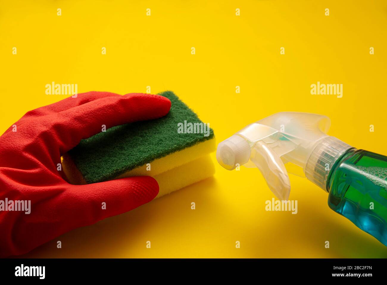 Hand in roten Gummihandschuh mit Schwamm und Flüssigwaschmittel in Sparrenflasche auf gelbem Hintergrund, Reinigungs- und Sanierungskonzept Stockfoto