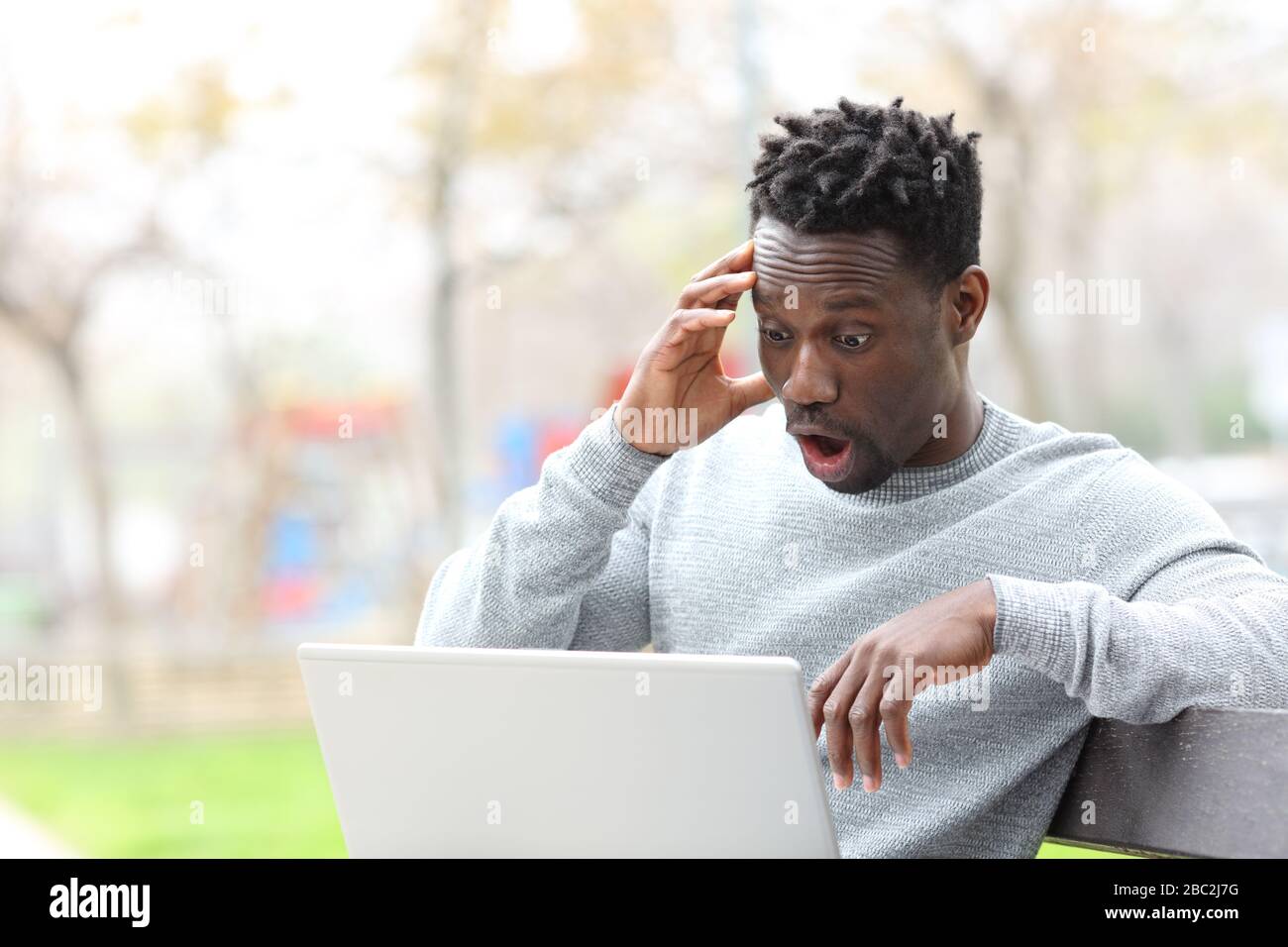 Staunender schwarzer Mann, der überraschende Nachrichten auf dem Laptop liest, der auf der Parkbank sitzt Stockfoto