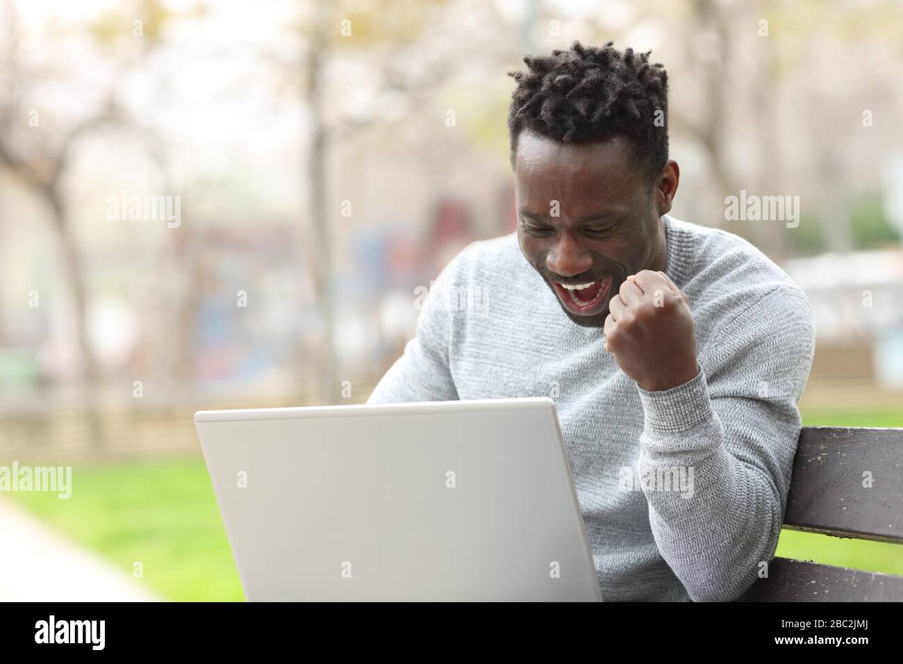 Aufgeregter schwarzer Mann, der die Überprüfung von Laptop-Inhalten feiert, die auf einer Bank in einem Park sitzen Stockfoto