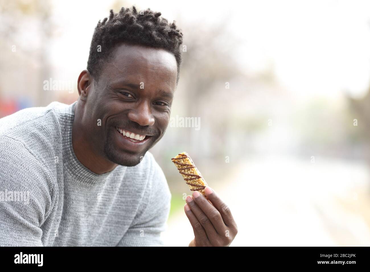 Fröhlicher schwarzer Mann, der die Granola-Bar zum Essen hält und die Kamera in einem Park mit Platz für Kopien betrachtet Stockfoto