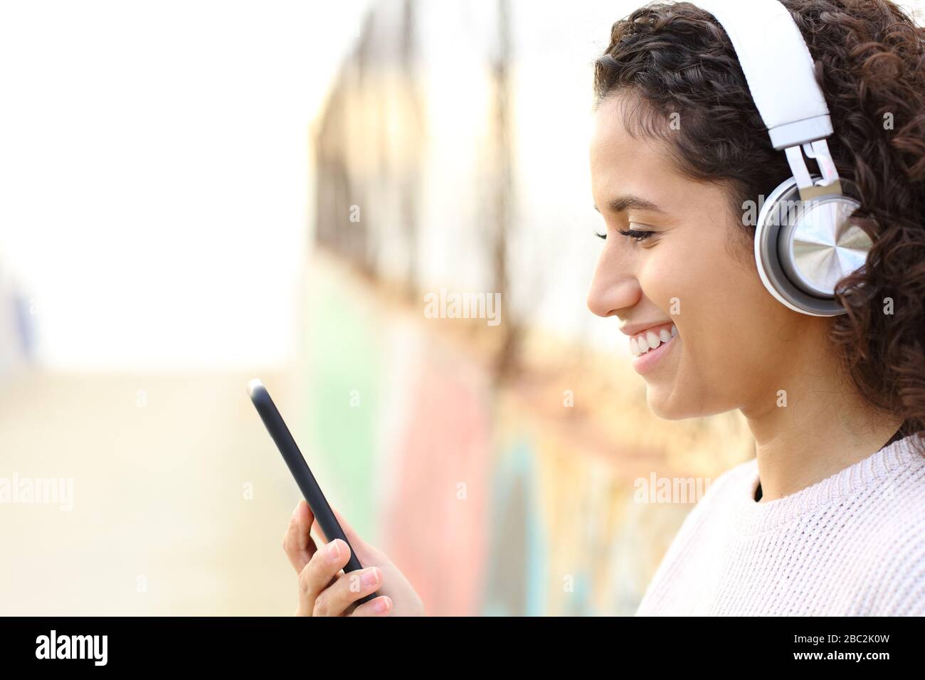 Seitenansicht Portrait eines glücklichen Latina-Mädchens, das Musik hört, Kopfhörer trägt und das Smartphone auf der Straße überprüft Stockfoto