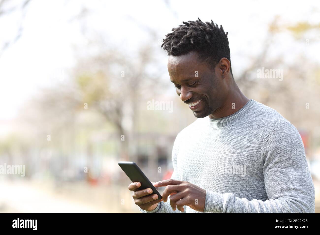 Glücklicher schwarzer Mann mit Smartphone, der einen sonnigen Tag in einem Park spaziert Stockfoto
