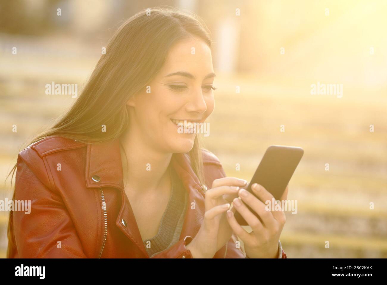Glückliche Frau, die mit einem Smartphone auf der Straße sitzt und ein warmes Licht hat Stockfoto