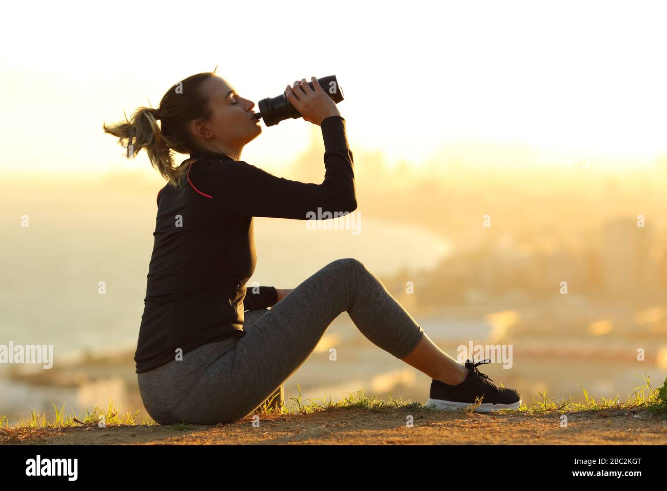 Seitenansicht Portrait eines ganzen Körpers einer Läuferin, die bei Sonnenuntergang am Stadtrand Trinkwasser aus einer Flasche stillt Stockfoto