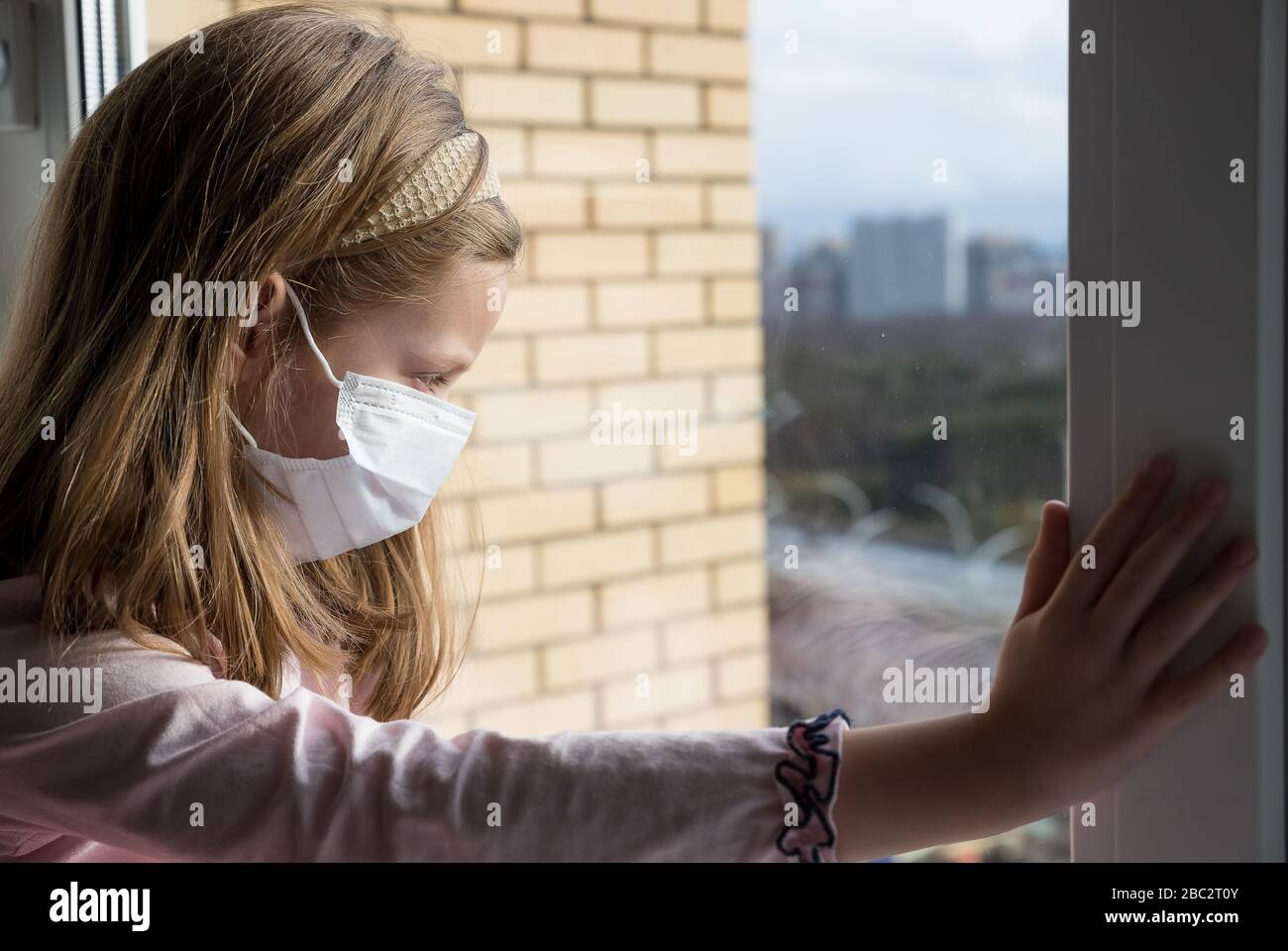 Ein Mädchen in einer Schutzmaske vor dem Virus auf ihrem Gesicht sitzt am Fenster, Blick auf die Straße.Prävention Epidemie.traurige Kind Mädchen in medizinische Maske durch die Stockfoto