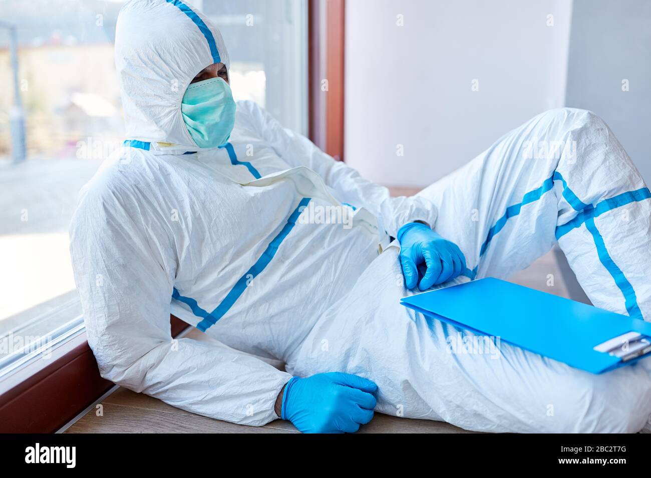 Doktor liegt in Schutzkleidung mit Stress und Burnout in der Klinik, die sich an Fenster bei der Covid-19-Coronavirus-Epidemie anlehnt Stockfoto