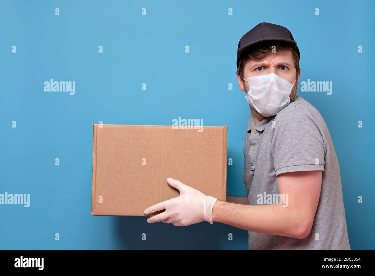 Kaukasischer junger Mann in medizinischer Maske, der ein Paket hält, das er dem Kunden gibt. Kein ansteckendes Lieferkonzept. Studio an blauer Wand gedreht. Stockfoto