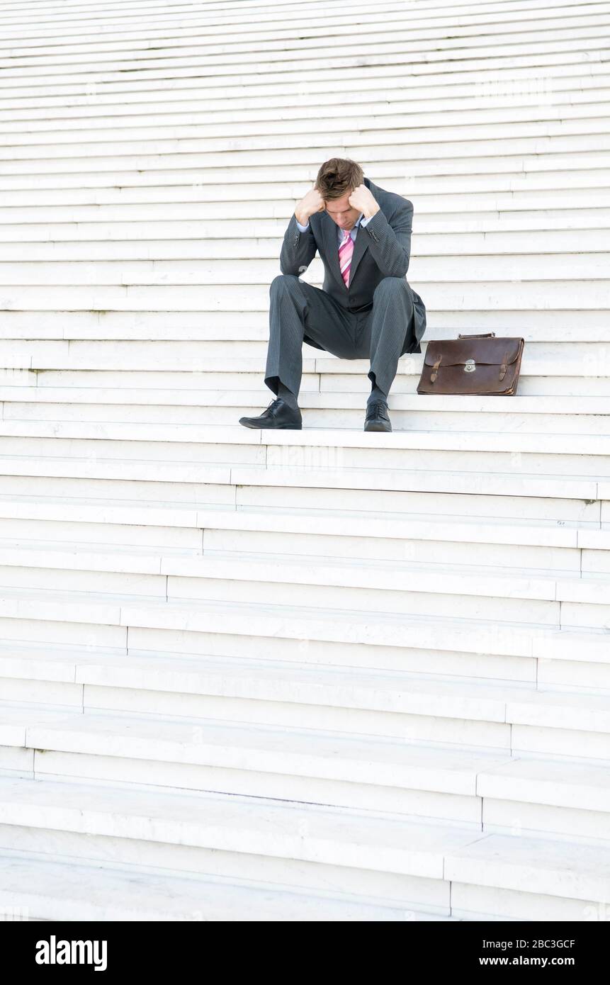 Depressiver Geschäftsmann sitzt draußen auf einer großen weißen Treppe, der seinen Kopf in den Händen hängt Stockfoto