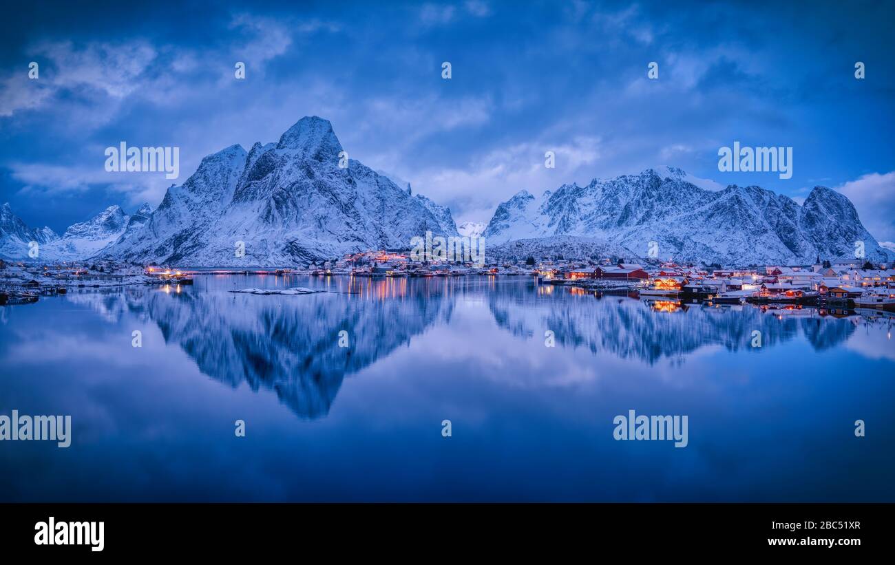 Die Abendlichter des Dorfes Reine, mit schneebedeckter Bergkette im Rücken, und seine Reflexion im ruhigen Wasser des Fjords. Stockfoto