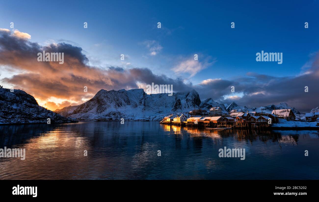 Am späten Nachmittag scheint Licht auf die Küstengebäude, mit Reflexionen im Wasser und schneebedeckten Bergen im Hintergrund. Stockfoto