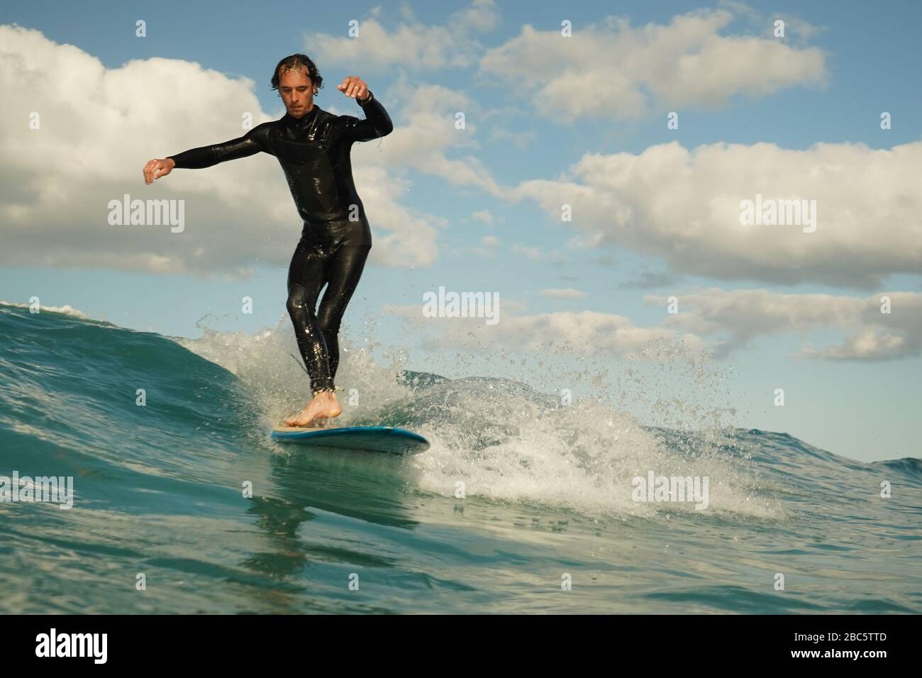 Ein junger männlicher Surfer in einem schwarzen Neoprenanzug reitet auf einer Bruchwelle mit weißem Wasser ein Longbord-Surfbrett. Sommertag mit blauem bewölktem Himmel. Stockfoto